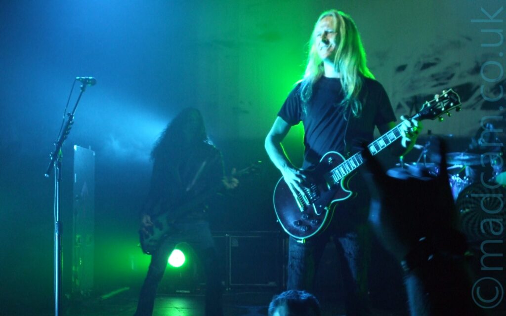 A man with long blonde hair, wearing a black t-shirt and jeans, and playing a black electric guitar standing on the right of the frame, with a microphone on a stand on the left. In the background, there is a person with long dark hair , wearing a leather jacket, and playing a black and silver bass guitar, standing on the left of the frame, shrouded in a haze of smoke and green and blue lights. A drum kit is visible in the background on the right, in front of a large backdrop where a video is being projected. The whole frame has a blue and green cast from all the stage lights.