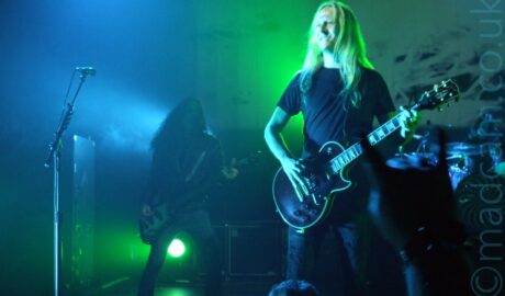 A man with long blonde hair, wearing a black t-shirt and jeans, and playing a black electric guitar standing on the right of the frame, with a microphone on a stand on the left. In the background, there is a person with long dark hair , wearing a leather jacket, and playing a black and silver bass guitar, standing on the left of the frame, shrouded in a haze of smoke and green and blue lights. A drum kit is visible in the background on the right, in front of a large backdrop where a video is being projected. The whole frame has a blue and green cast from all the stage lights.
