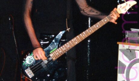 A tall, long-limbed man with short, slightly spiky ginger hair, wearing a black tank top under a long-sleeved black mesh top that shows off the tattoos on his arms, and black trousers, playing a black bass guitar, looking down at his instrument. Black walls and ceiling fill most of the background, with a large, partially rolled grey and white poster stretching across the frame from the left, and a green stage light shining over his right shoulder.