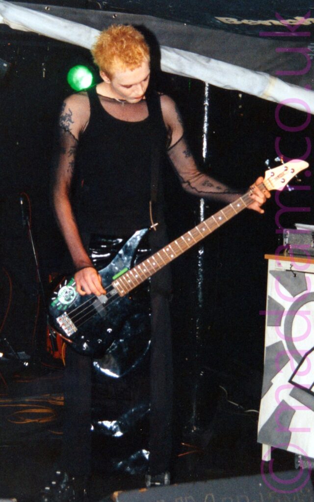 A tall, long-limbed man with short, slightly spiky ginger hair, wearing a black tank top under a long-sleeved black mesh top that shows off the tattoos on his arms, and black trousers, playing a black bass guitar, looking down at his instrument. Black walls and ceiling fill most of the background, with a large, partially rolled grey and white poster stretching across the frame from the left, and a green stage light shining over his right shoulder.