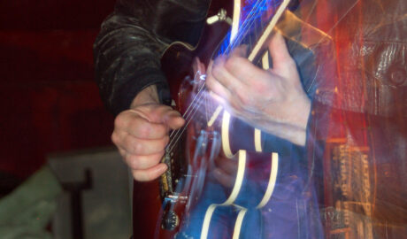 Close-up of a polished brown electric guitar with white piping, being played by a man with a hairy chin and wearing a battered black leather jacket and dark blue jeans. A flash from the camera has frozen him in place, while a long exposure and a snap zoom out results in a secondary, smaller image of him in the middle of the frame, with streaks of blue, white, and red lights. Red walls fill the rest of the frame.