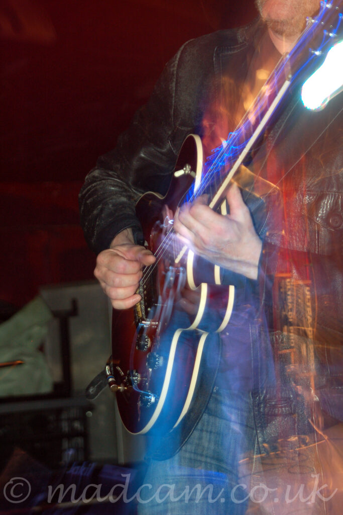 Close-up of a polished brown electric guitar with white piping, being played by a man with a hairy chin and wearing a battered black leather jacket and dark blue jeans. A flash from the camera has frozen him in place, while a long exposure and a snap zoom out results in a secondary, smaller image of him in the middle of the frame, with streaks of blue, white, and red lights. Red walls fill the rest of the frame.